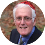 Elderly man with glasses, wearing a red hat and black suit, smiling outdoors.