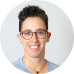 Person with short curly hair and glasses, wearing a light blue top, smiles against a neutral background.