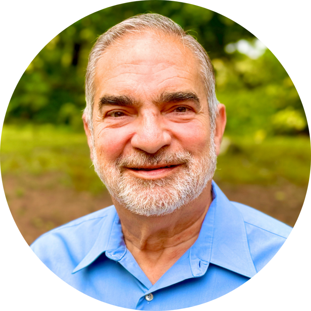 Smiling man with a beard wearing a blue shirt, standing outdoors with trees in the background.