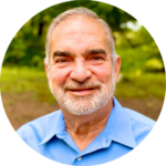 Smiling man with a beard wearing a blue shirt, standing outdoors with trees in the background.