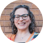 Person with glasses and curly hair smiling, standing against a brick wall.