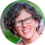 Smiling person with curly hair and glasses, outdoors with greenery in the background.