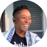 Rabbi Sandra Lawson smiling warmly, wearing a black t-shirt and headphones, standing outdoors near a window.