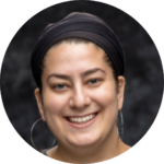 Person with a black headband and hoop earrings smiling against a dark, blurred background.