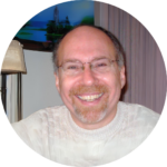 Smiling man with glasses and a beard, wearing a white sweater, indoors with a lamp and painting in the background.