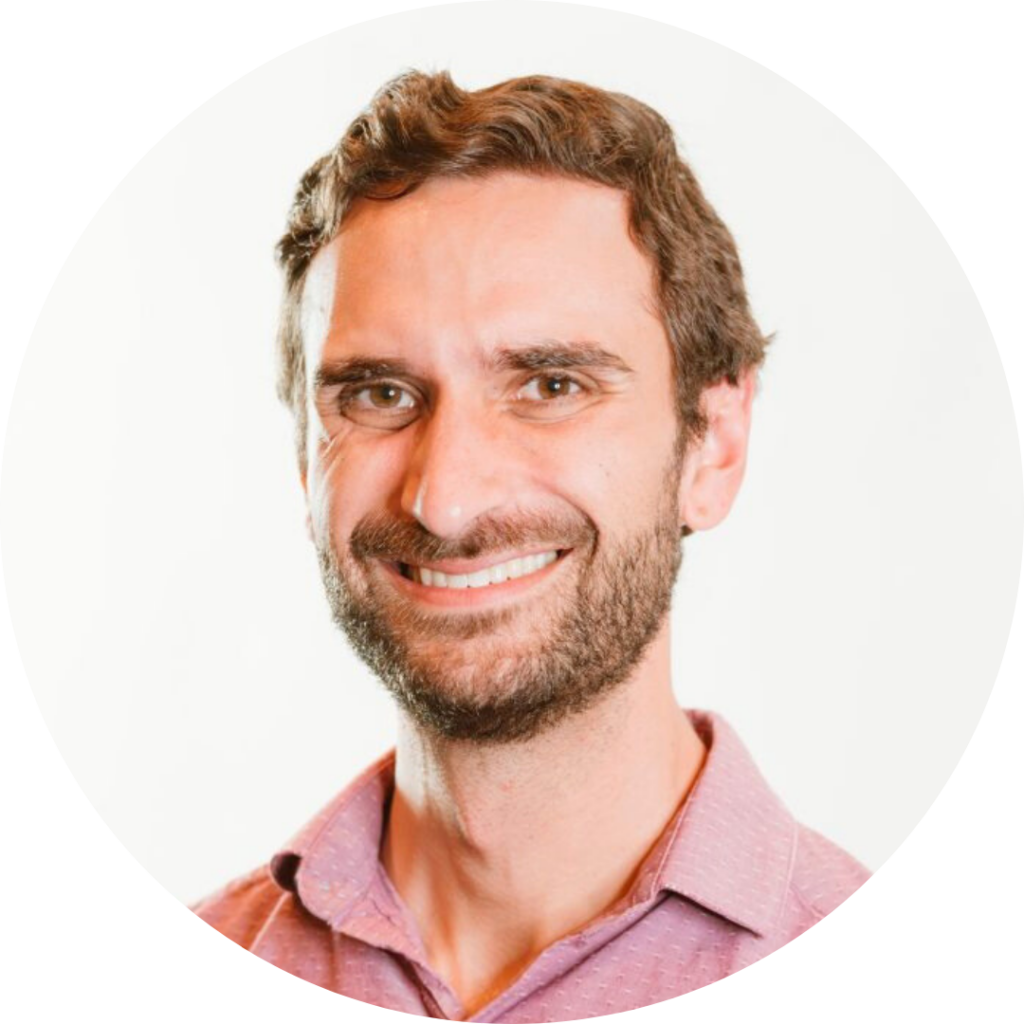 Man with short brown hair and a beard, wearing a pink shirt, smiling against a white background.