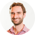 Man with short brown hair and a beard, wearing a pink shirt, smiling against a white background.