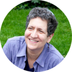 A person with short curly hair and a blue shirt smiling outdoors on grass.