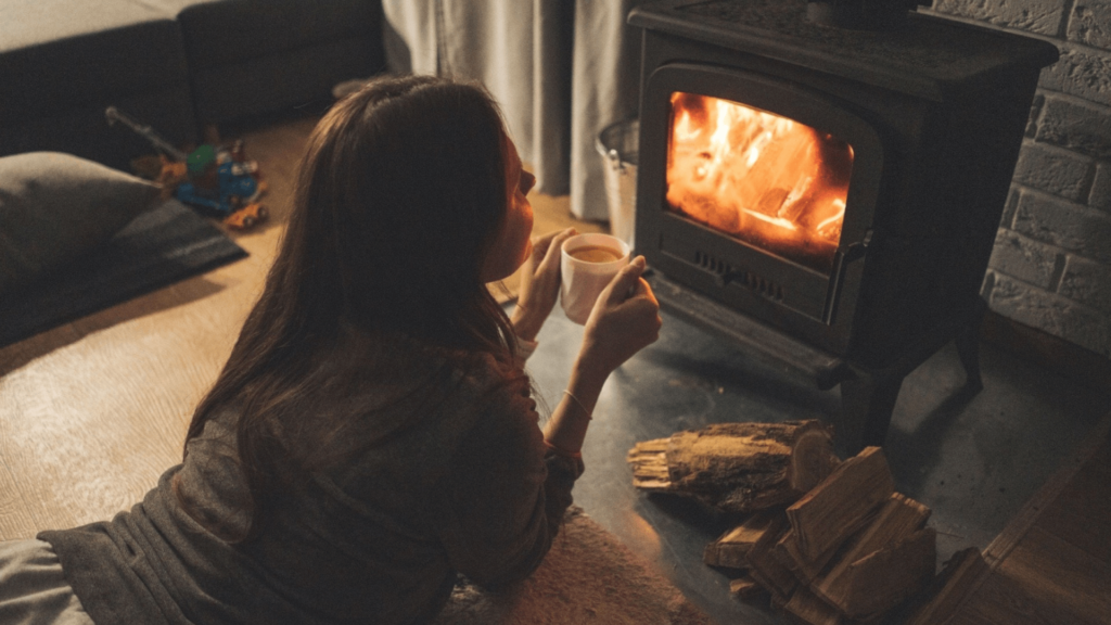 A person relaxes by a wood-burning stove, holding a mug, with firewood nearby in a cozy, dimly lit room.