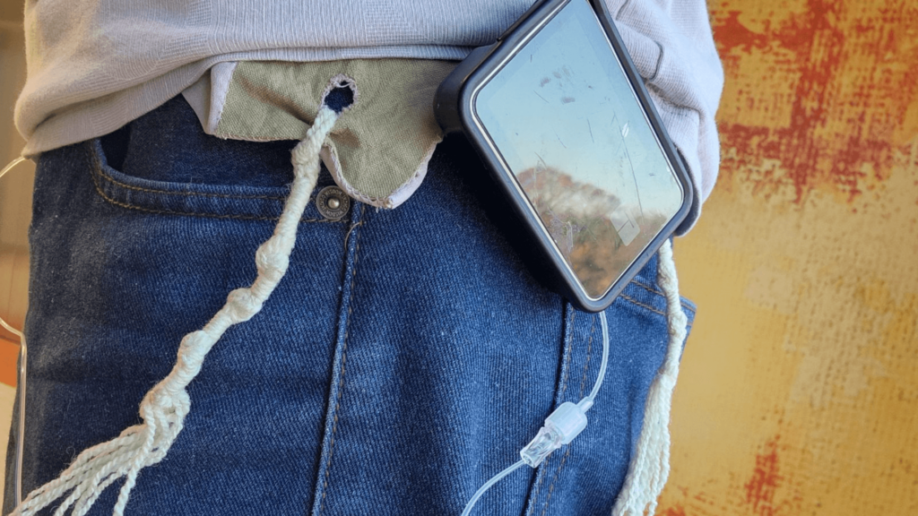 A phone in a gray case on the waist of a person's jeans with medical tubing, next to talit tassels