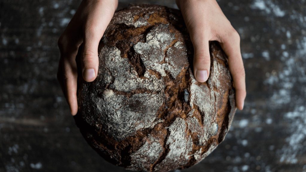 Hands holding a round, rustic loaf of bread with a dark, crusty surface.