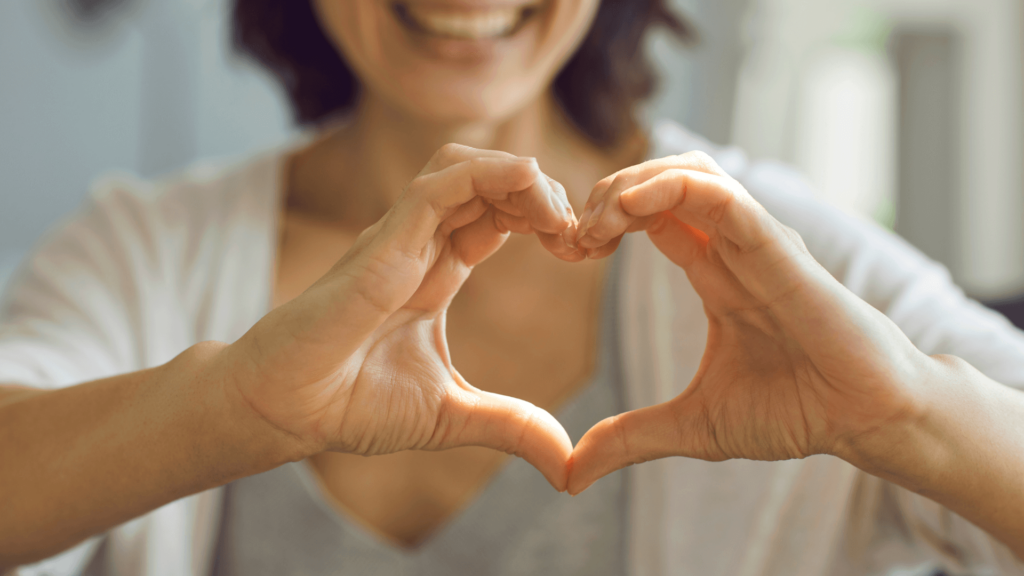 Person smiling and forming a heart shape with their hands, wearing a gray shirt and white cardigan.