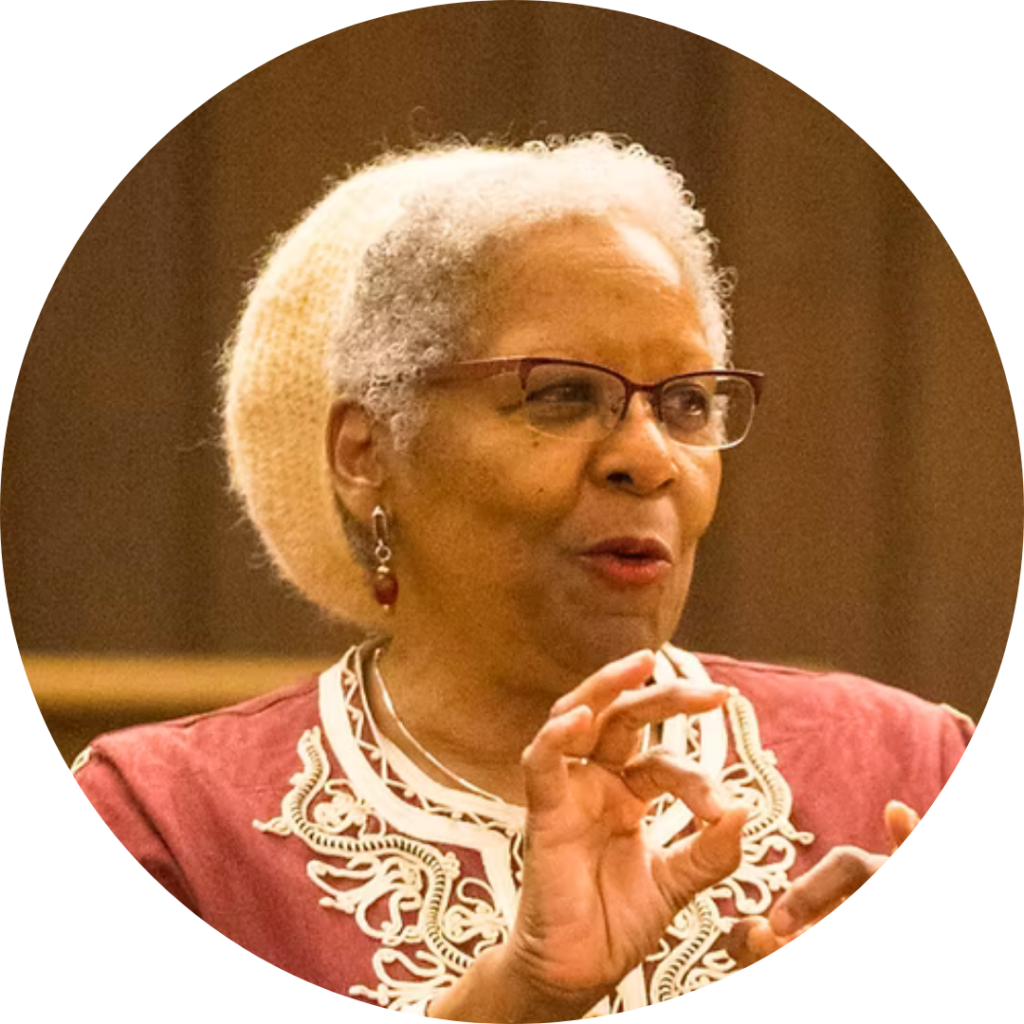 Elderly woman with short gray hair, wearing glasses and a patterned top, gesturing with her hand.