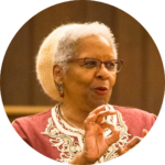 Elderly woman with short gray hair, wearing glasses and a patterned top, gesturing with her hand.