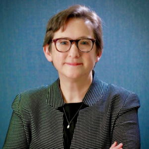 Person with glasses and short hair, wearing a black and white patterned suit, standing against a blue background.