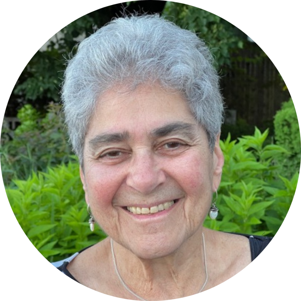 An older person with short gray hair smiles, standing outdoors with green foliage in the background.