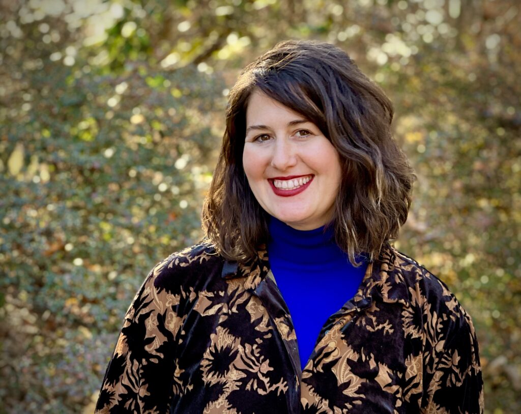 Alana Krivo-Jaufman, wearing a patterned jacket and blue turtleneck stands outdoors with leafy background.