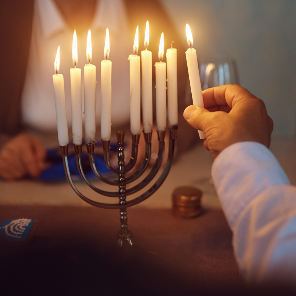 A person lighting a menorah candle, with another person sitting in the background at a table.