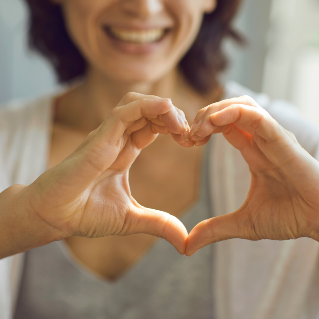 A person making a heart shape with their hands, smiling in the background.