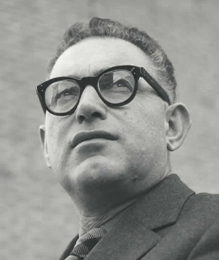 A person in glasses looks into the distance against a plain background.