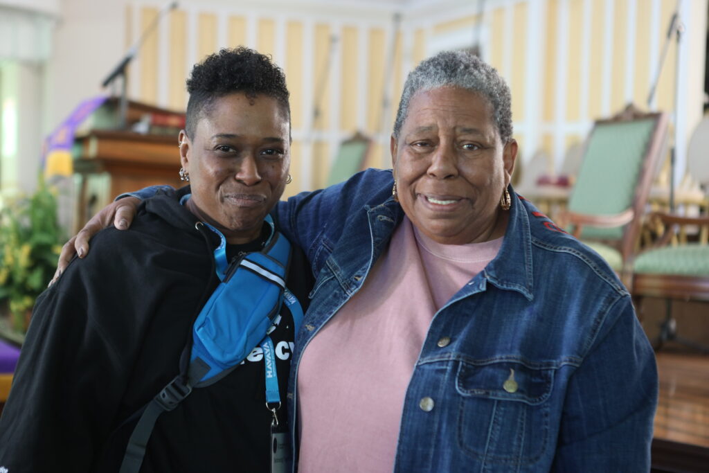 Joanne Bland and Rabbi Sandra Lawson smiling and posing together, standing in a room with chairs and musical instruments in the background.