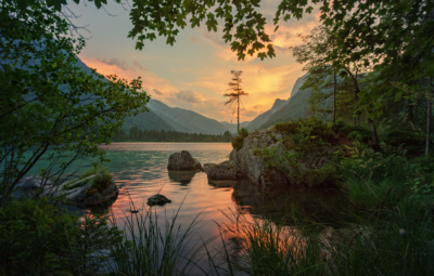Serene lake at sunset, framed by trees and mountains, with vibrant orange and pink sky reflecting on the water.