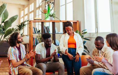 A group of people sitting in a circle, clapping and smiling, in a bright room with plants and bookshelves.