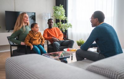 A family of three sits on a sofa, talking with a man in a living room with a TV and plants.