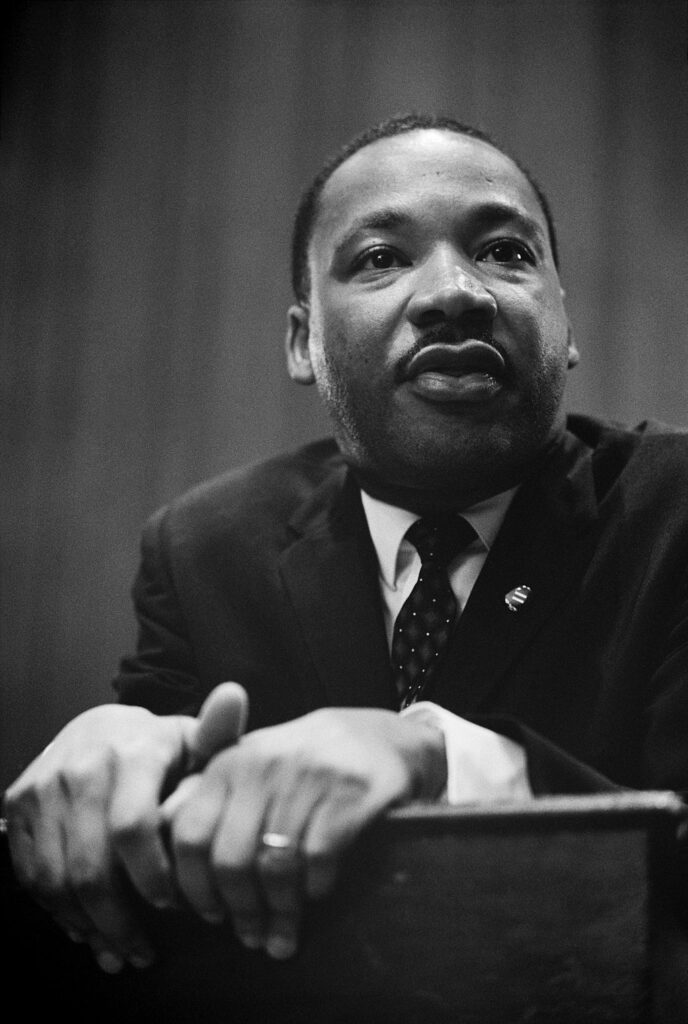 Martin Luther King, Jr. in a suit speaks while leaning on a podium, his hands resting gently.
