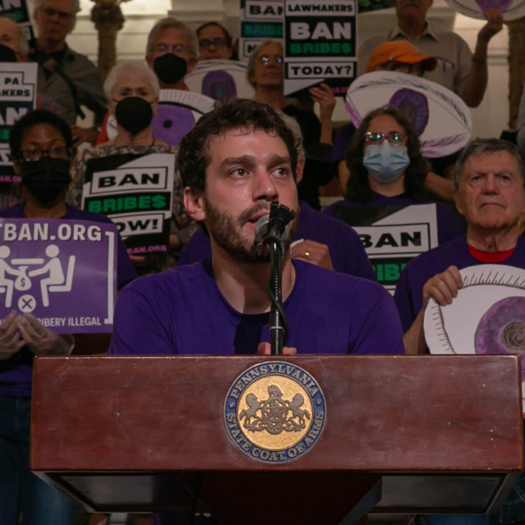 A person speaks at a podium, surrounded by a crowd holding signs advocating to ban bribery.