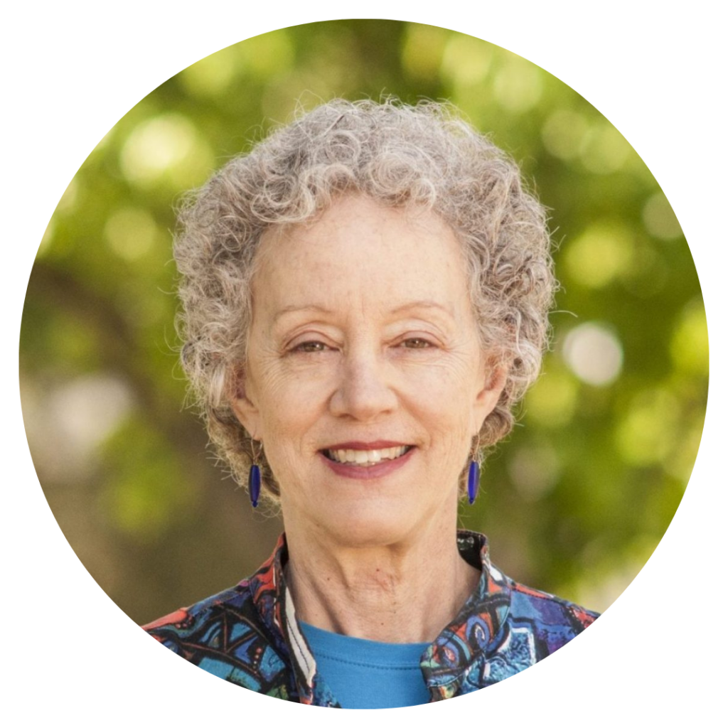 Person with curly gray hair, wearing colorful jacket and blue earrings, smiling against a leafy background.