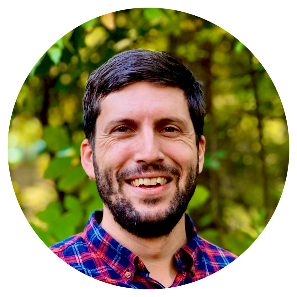 Smiling man with a beard wearing a plaid shirt, standing in front of a leafy green background.