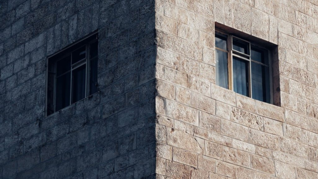 Corner of a stone building with two windows, one on each visible side, under a clear sky.