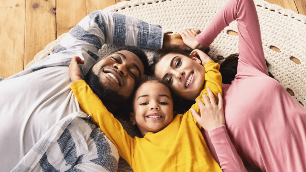A happy multiracial family of a father, mother, and daughter lying on a rug, smiling.