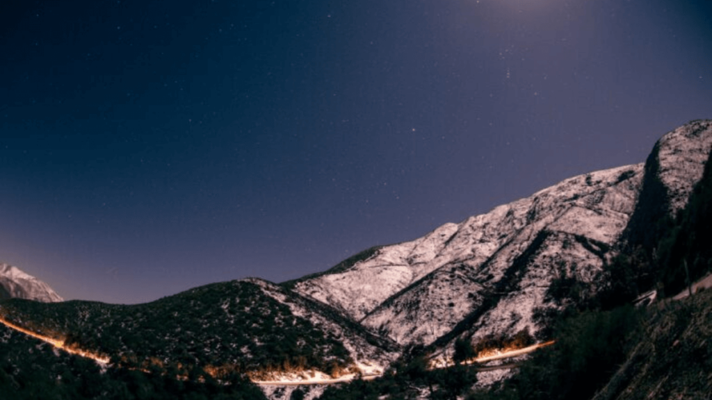 Snow-covered mountains under a starry night sky, with a winding road lit by car lights below.