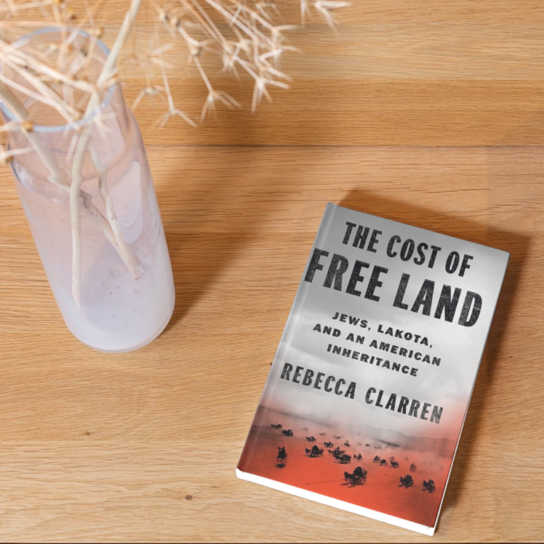 Book titled The Cost of Free Land by Rebecca Clarren on a wooden table next to a vase with dried plants.