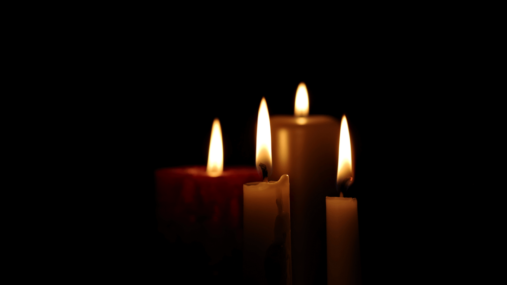 Four lit candles in a dark setting, with flames softly illuminating their surroundings.