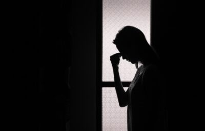 Silhouette of a person standing in front of a window, holding their head with one hand, appearing pensive.