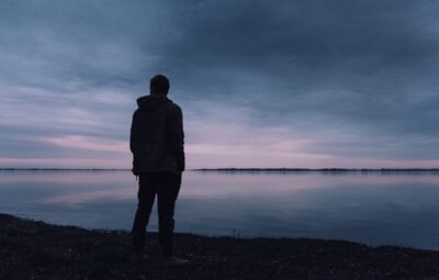 Silhouette of a person standing by a calm lake during a cloudy sunset, with soft pink and blue hues in the sky.