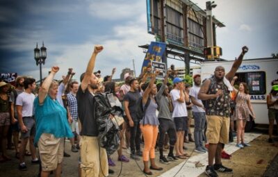 A diverse group of people hold up their fists and signs in protest on a street with overcast skies.