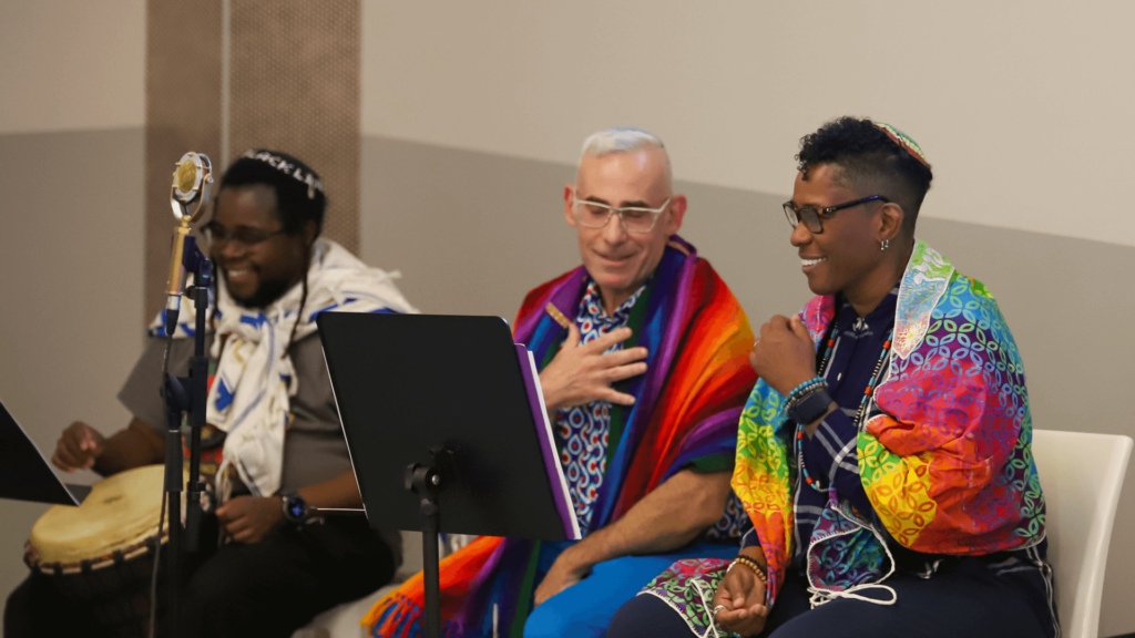 Three people, draped in colorful shawls, sit together with one playing a drum, smiling and engaged in conversation.