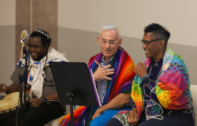 Three people, draped in colorful shawls, sit together with one playing a drum, smiling and engaged in conversation.