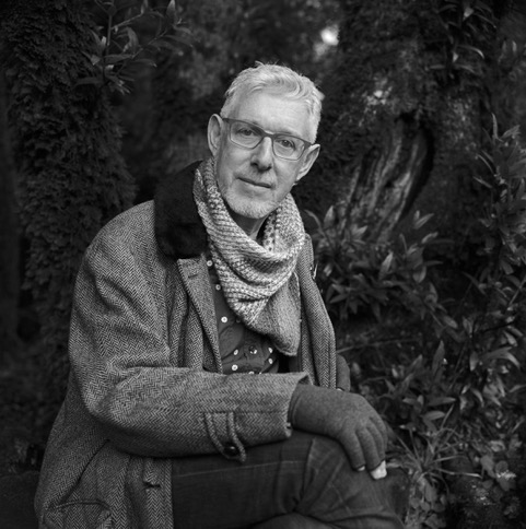 Man with glasses and scarf sits outdoors in a forest, wearing a coat and gloves, looking at the camera.
