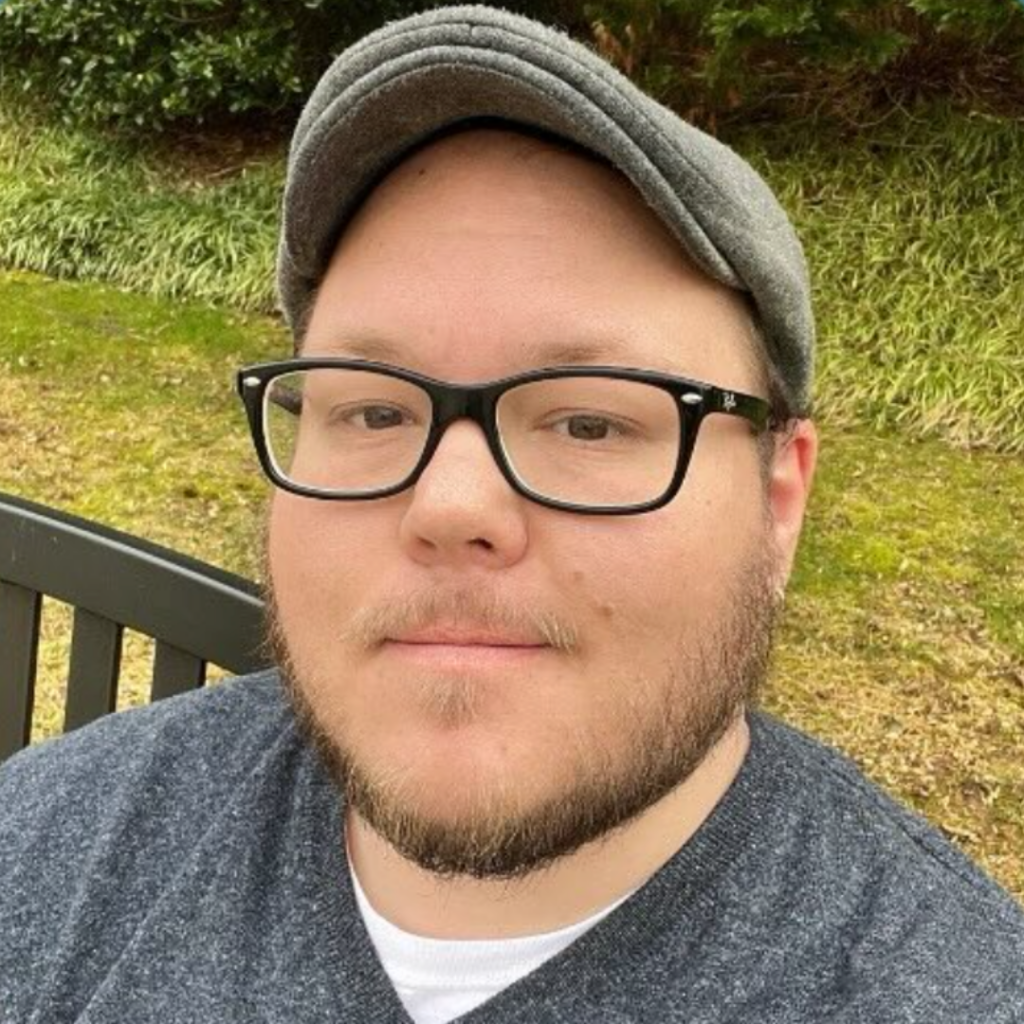 Person with a beard, glasses, and a hat sitting on a bench outdoors, with greenery in the background.