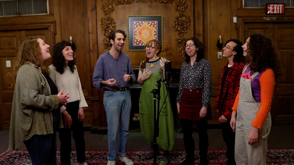 Seven people stand around a microphone in a room, singing and smiling warmly.