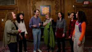 Seven people stand around a microphone in a room, singing and smiling warmly.