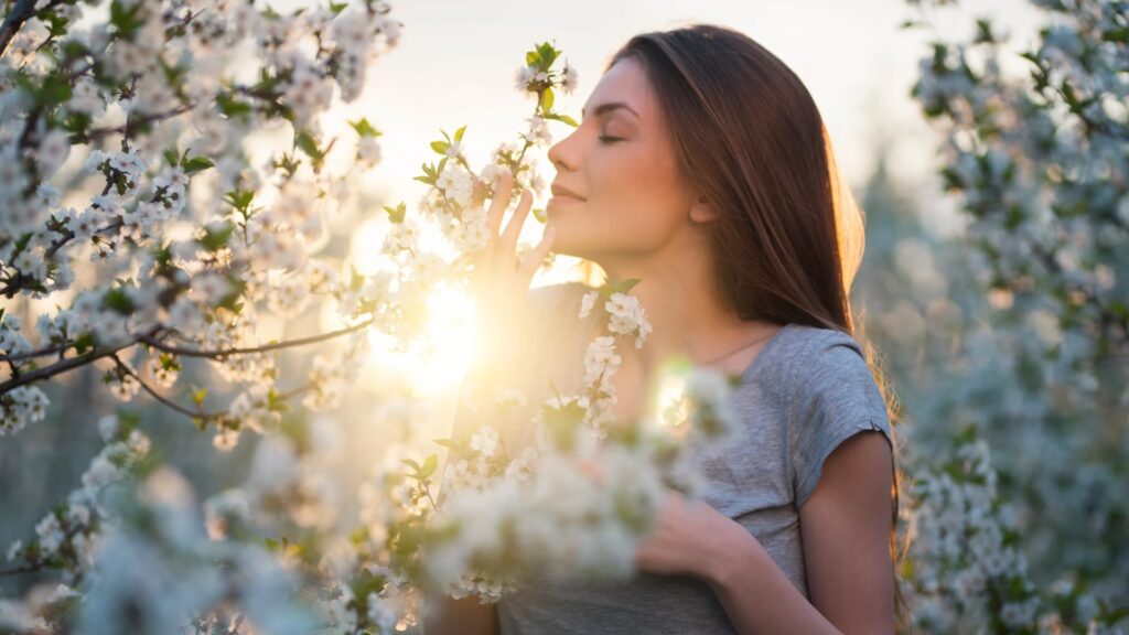 Woman in gray shirt smells blooming flowers on a sunny day, with sunlight shining through the blossoms.