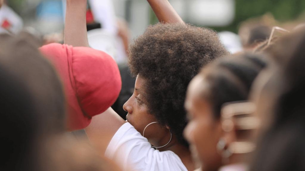 Person with afro and hoop earrings raises fist among a group, wearing a white shirt.