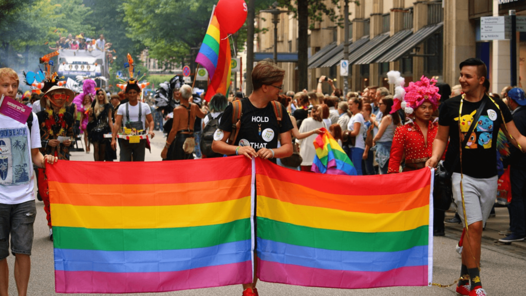People carrying a rainbow flag at a lively pride parade with colorful outfits and balloons.