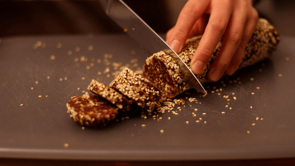 A hand slicing a sesame seed-covered roll on a cutting board.